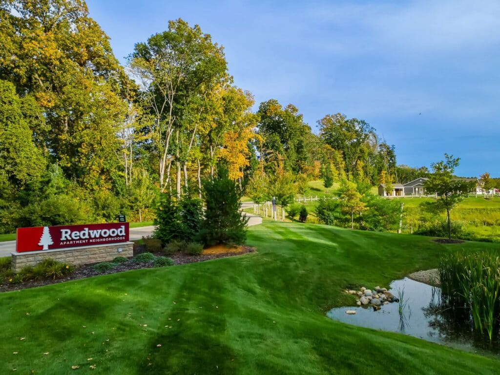 A landscaped green lawn with a small pond, trees, and a red sign that reads "Redwood Apartment Neighborhoods" on the left side. Buildings are visible in the background.