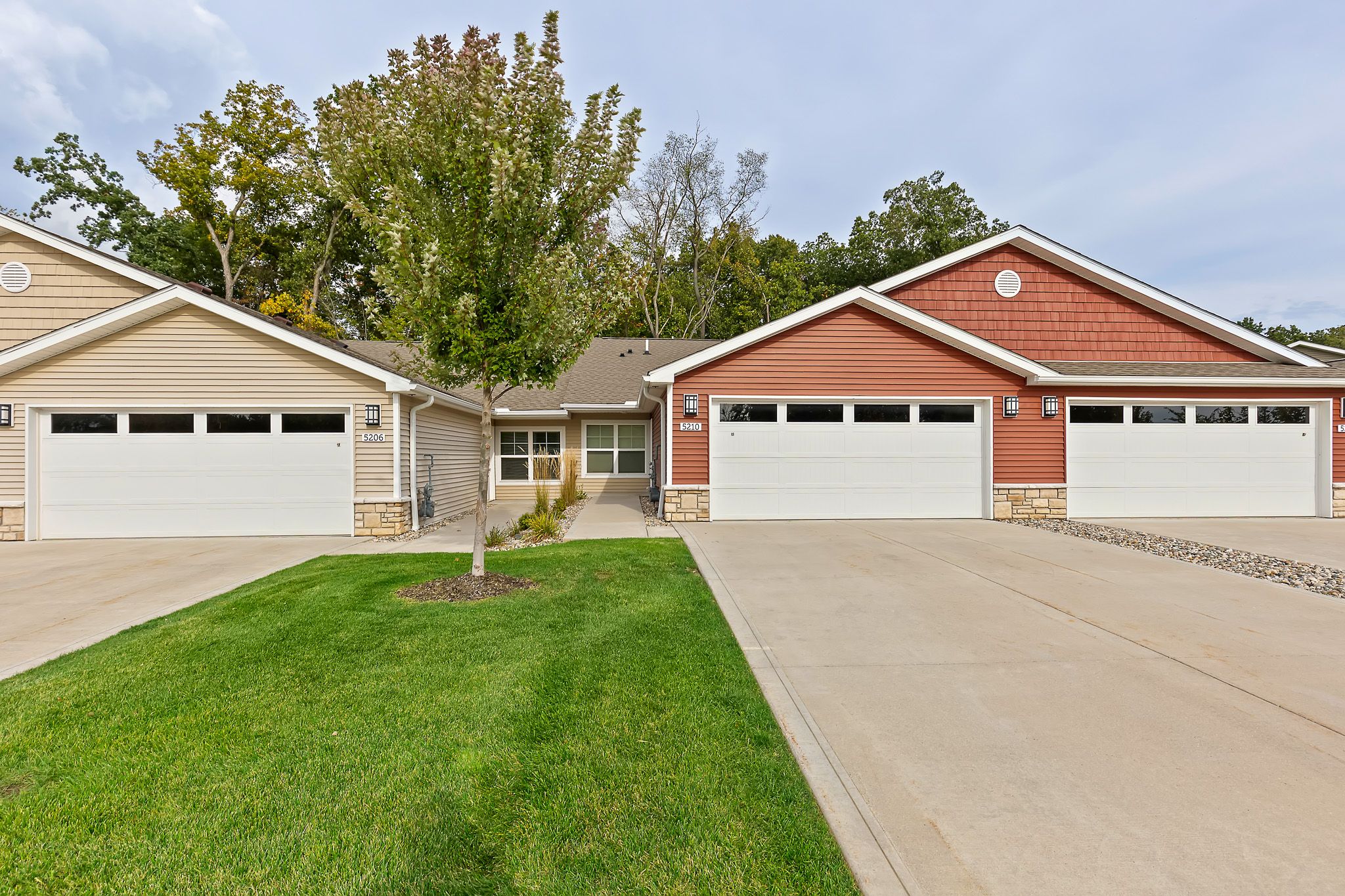A row of townhouses with beige and red siding, each featuring double garages, concrete driveways, and a small green lawn with a young tree in front.