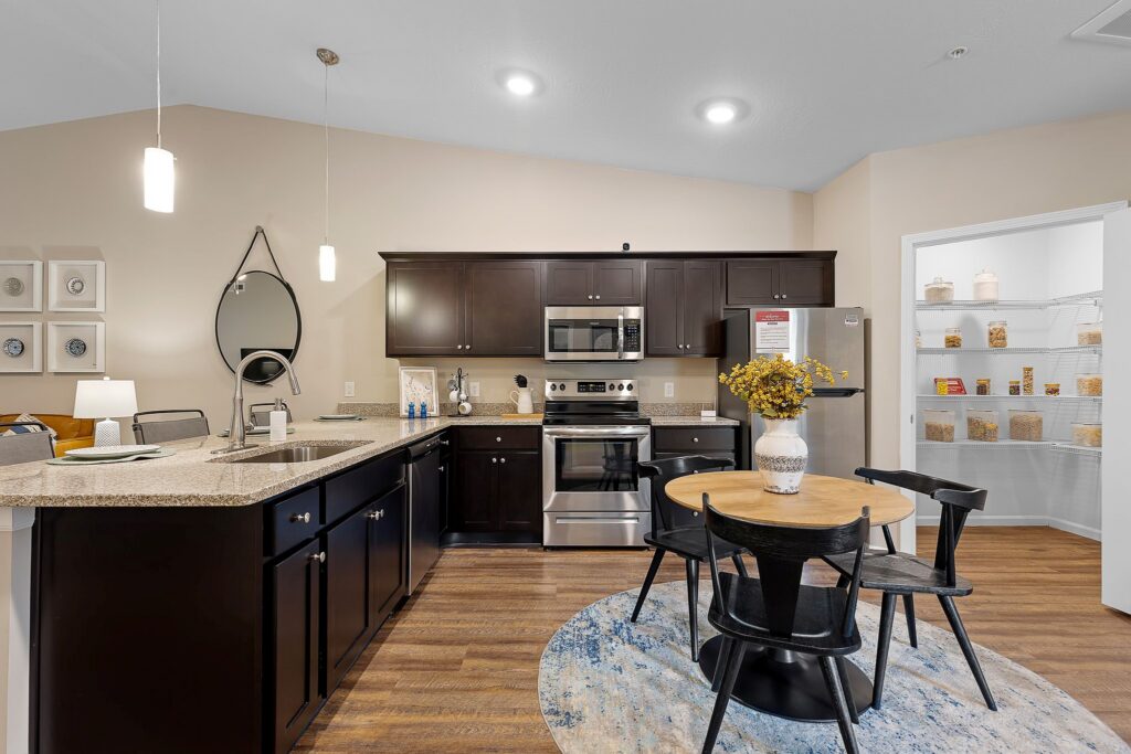 Modern kitchen with dark cabinets, stainless steel appliances, round dining table with four chairs, and a pantry stocked with food items in the background.