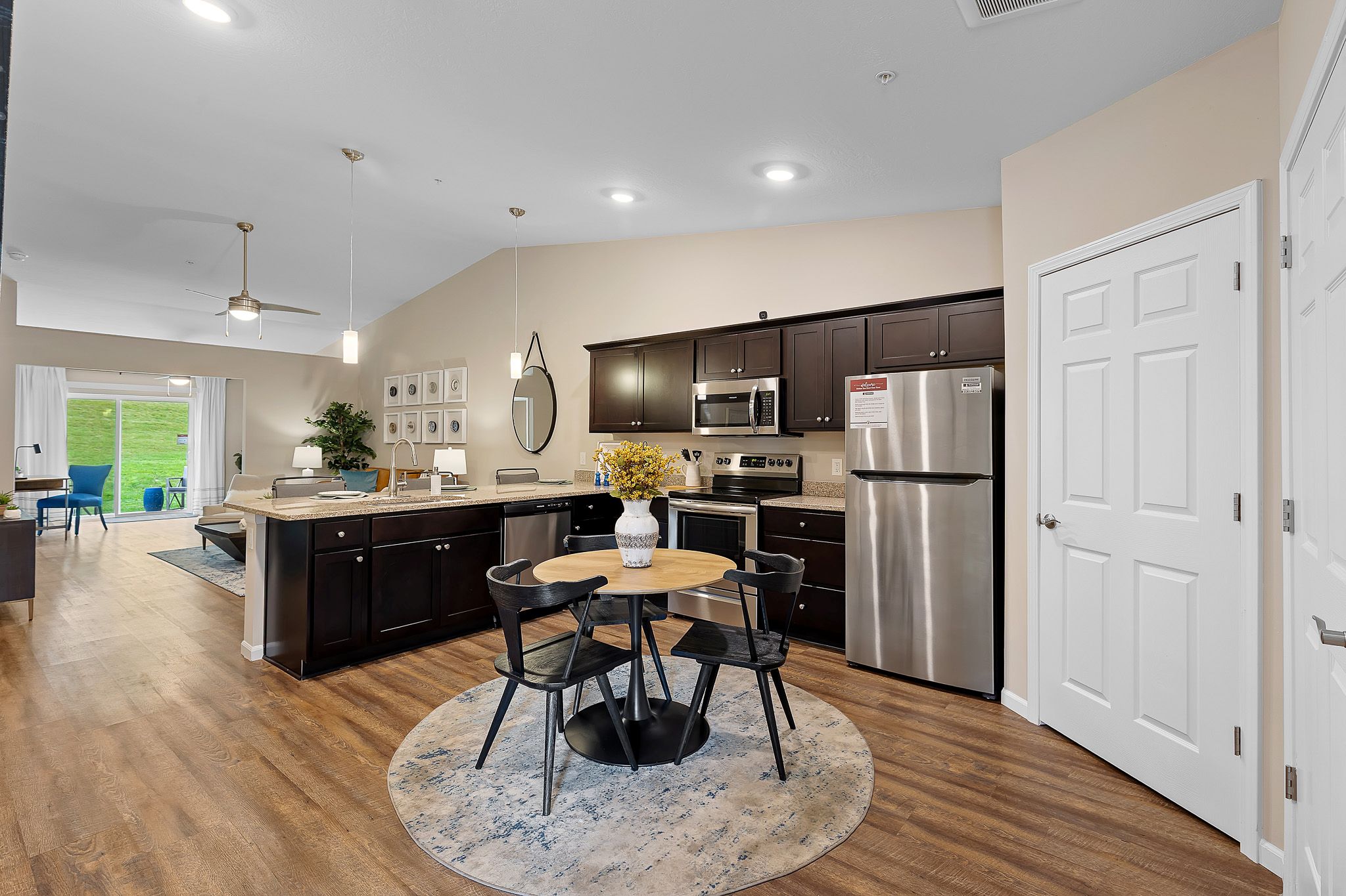 Modern kitchen with dark cabinets, stainless steel appliances, a round dining table with four chairs, and a vase of yellow flowers on a rug. Open layout with a view of a living area and patio.