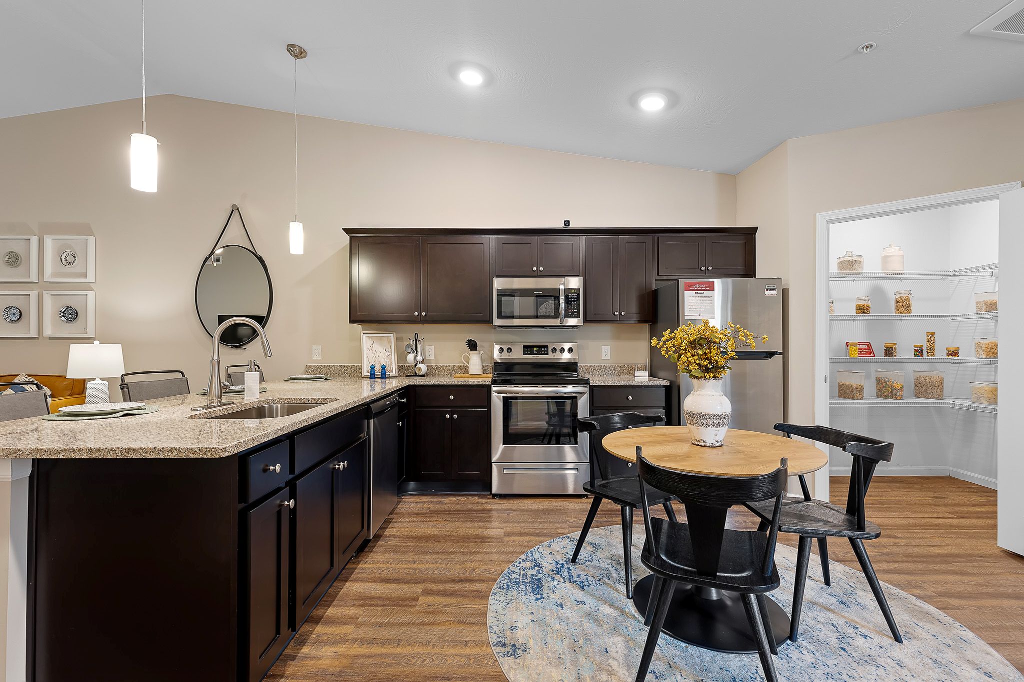 Modern kitchen with dark cabinets, stainless steel appliances, round dining table with four chairs, and a pantry stocked with food items in the background.