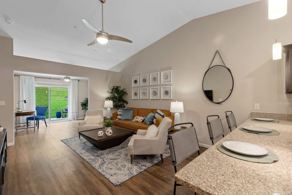 Modern living room with a brown sofa, two white chairs, a coffee table, and a patterned rug beside an open kitchen with a granite counter and bar stools; sliding doors lead outside.