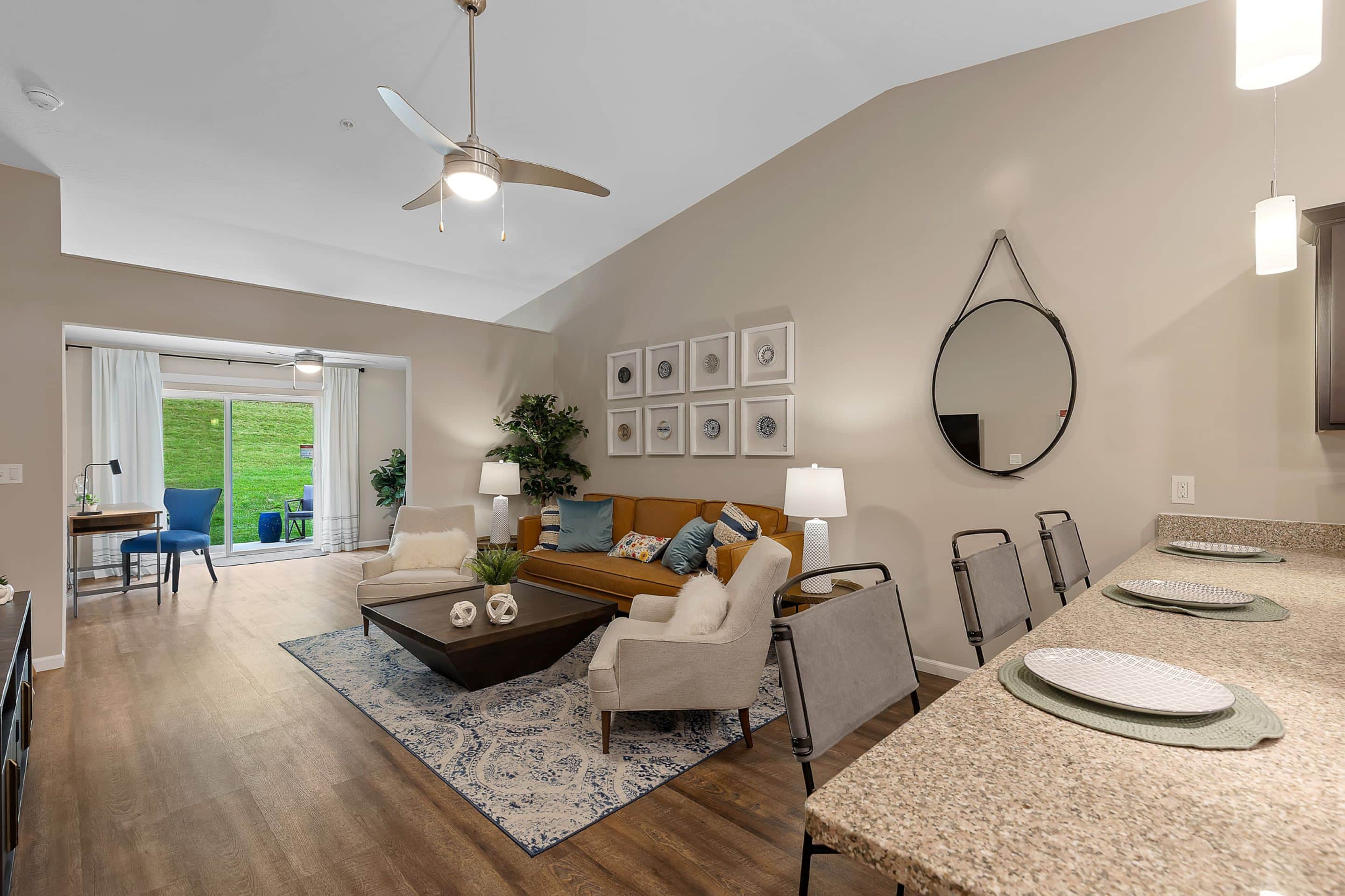 Modern living room with a brown sofa, two white chairs, a coffee table, and a patterned rug beside an open kitchen with a granite counter and bar stools; sliding doors lead outside.