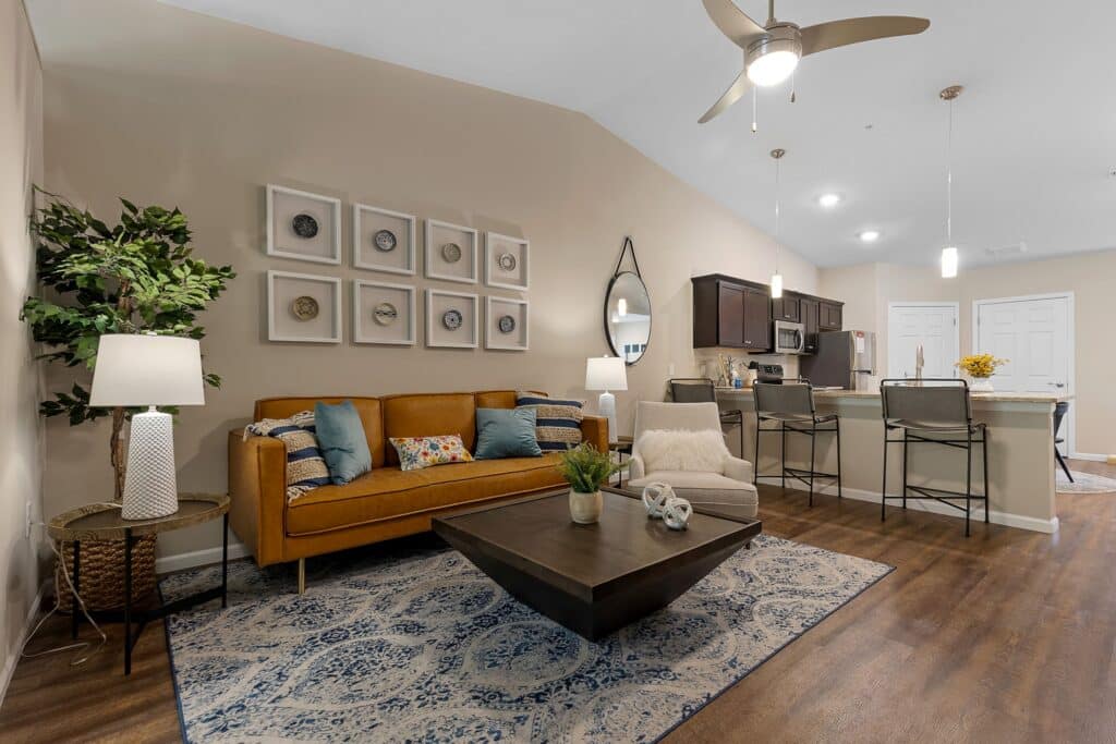 A modern living room with a brown sofa, armchair, coffee table, and decorative art on the wall; in the background, an open kitchen with bar stools and pendant lights.