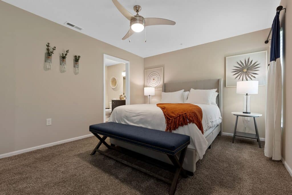 Modern bedroom with beige walls, queen bed with white linens and an orange throw, blue bench at foot, two side tables with lamps, wall art, and an open doorway to a bathroom.
