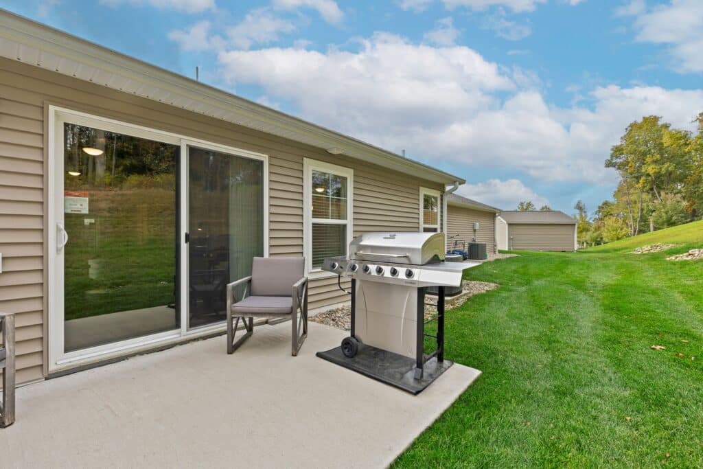 A small concrete patio with a grill and two chairs outside a beige house, with a sliding glass door and green lawn under a partly cloudy sky.