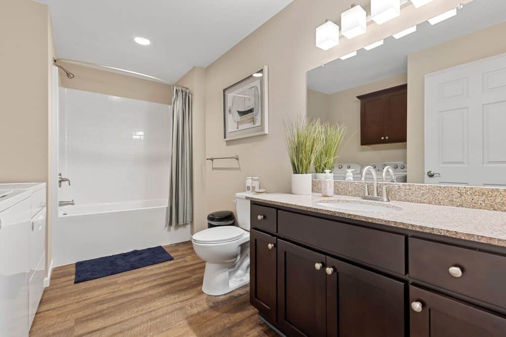 Modern bathroom with dark wood vanity, granite countertop, a toilet, bathtub with shower curtain, and laundry machines visible in the background.
