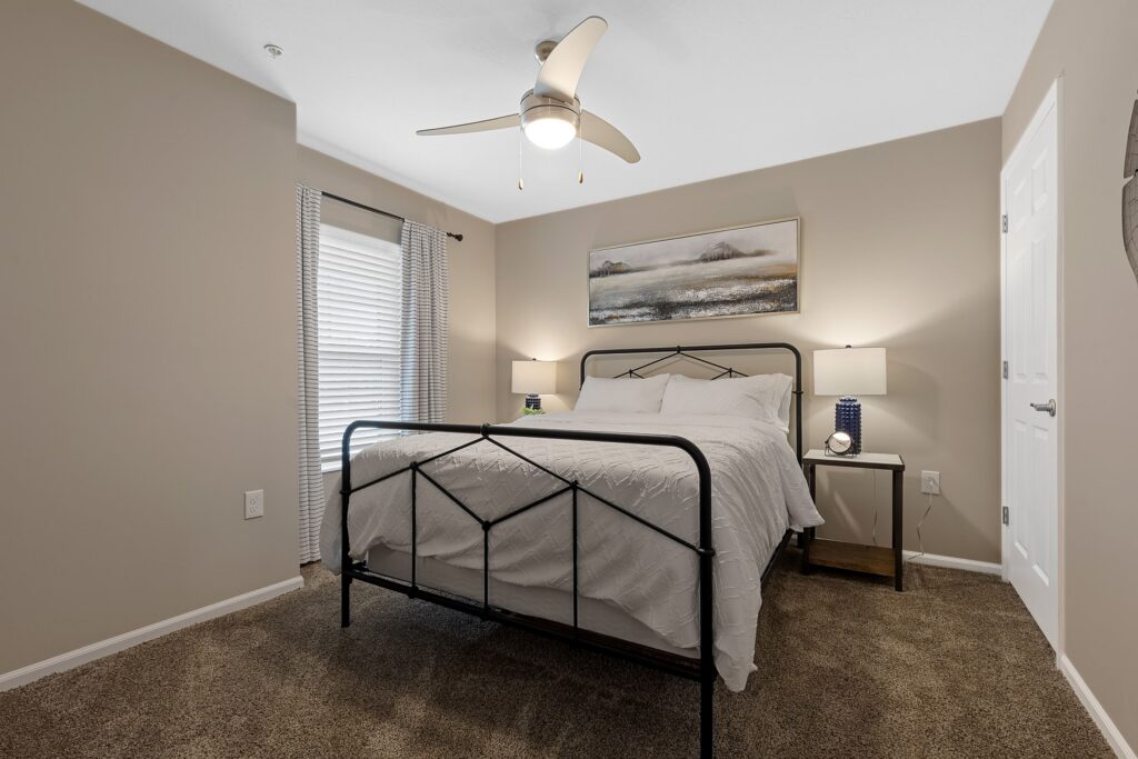 A neatly made bed with white linens and black metal frame is centered in a carpeted bedroom with beige walls, two nightstands, lamps, and a ceiling fan.