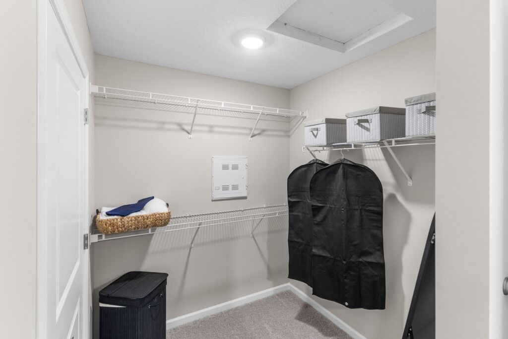 A walk-in closet with wire shelves, storage bins, a laundry basket, a basket with towels, and two garment bags hanging on the side.