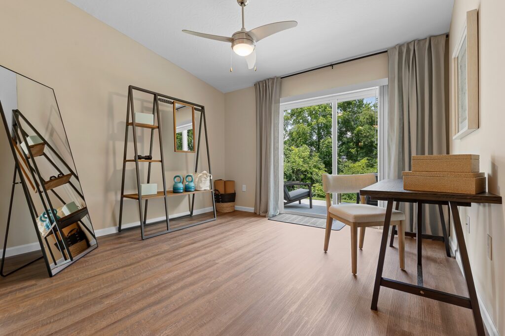 A minimalist room with wood flooring, a desk and chair, shelving, a floor mirror, and large window doors overlooking greenery.