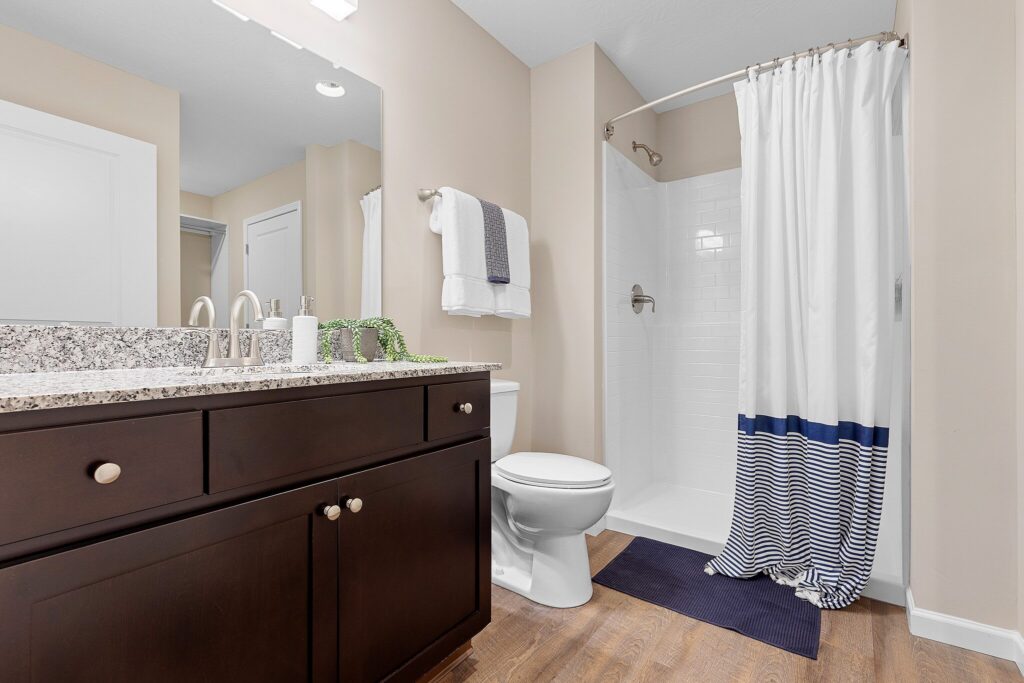 Modern bathroom with a granite countertop, dark wood vanity, white toilet, walk-in shower with white and blue striped curtain, and towels neatly arranged on a rack.