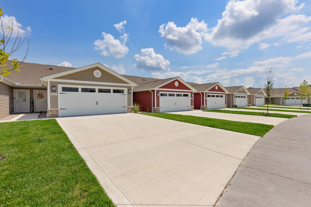 A row of modern single-story houses with attached garages and wide driveways on a sunny day, under a blue sky with scattered clouds.
