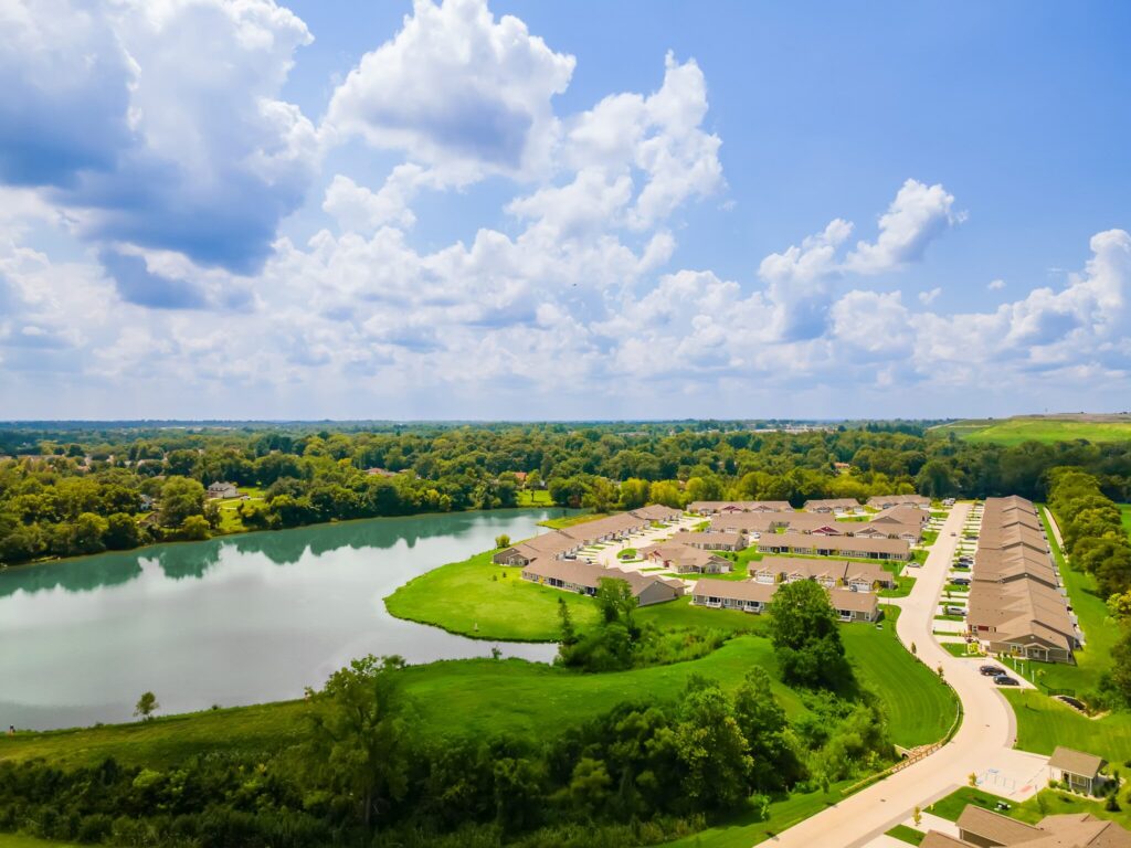 A suburban neighborhood with rows of houses is situated next to a lake, surrounded by green trees and grass under a partly cloudy sky.