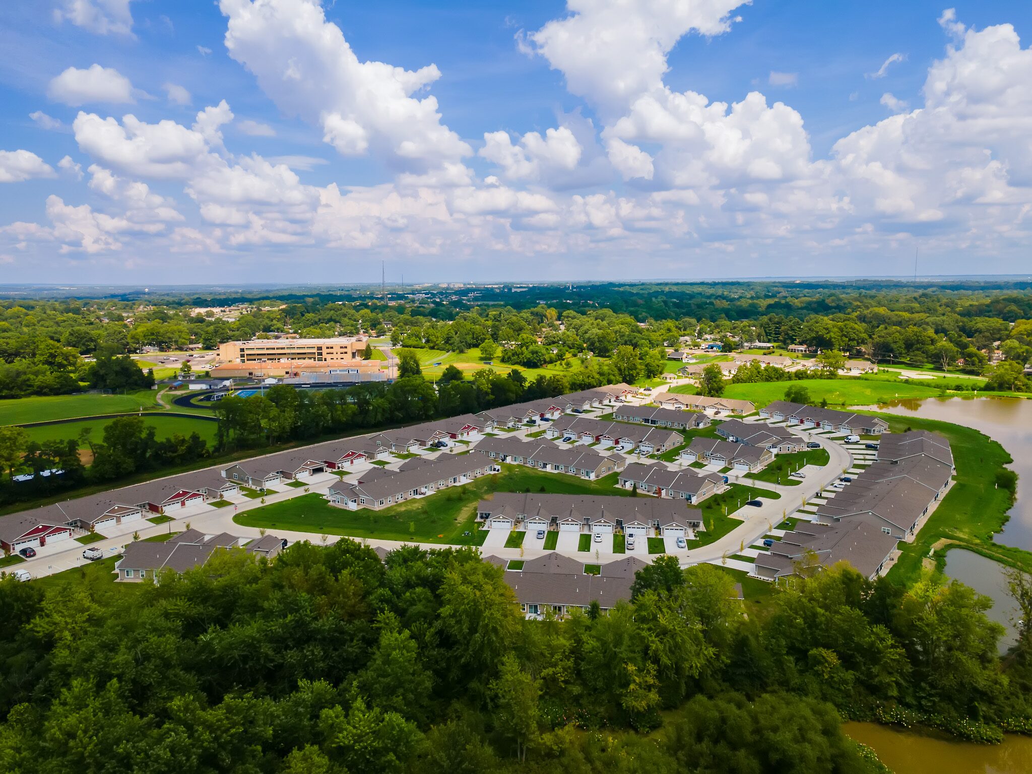 Aerial view of a suburban residential neighborhood with rows of houses, surrounding greenery, a body of water, and a large building in the background under a partly cloudy sky.