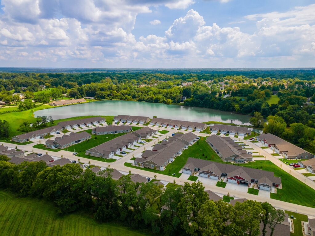 Aerial view of a suburban neighborhood with rows of houses, green lawns, a nearby pond, and surrounding trees under a partly cloudy sky.