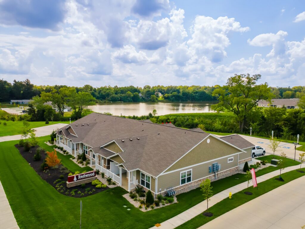 A single-story apartment complex with manicured lawns sits near a lake, surrounded by trees under a partly cloudy sky.