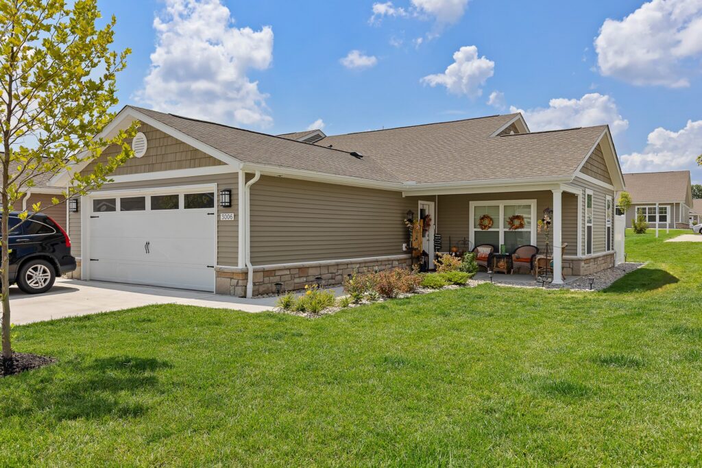 Single-story suburban house with beige siding, attached two-car garage, front porch with chairs, and a well-maintained lawn under a partly cloudy sky.