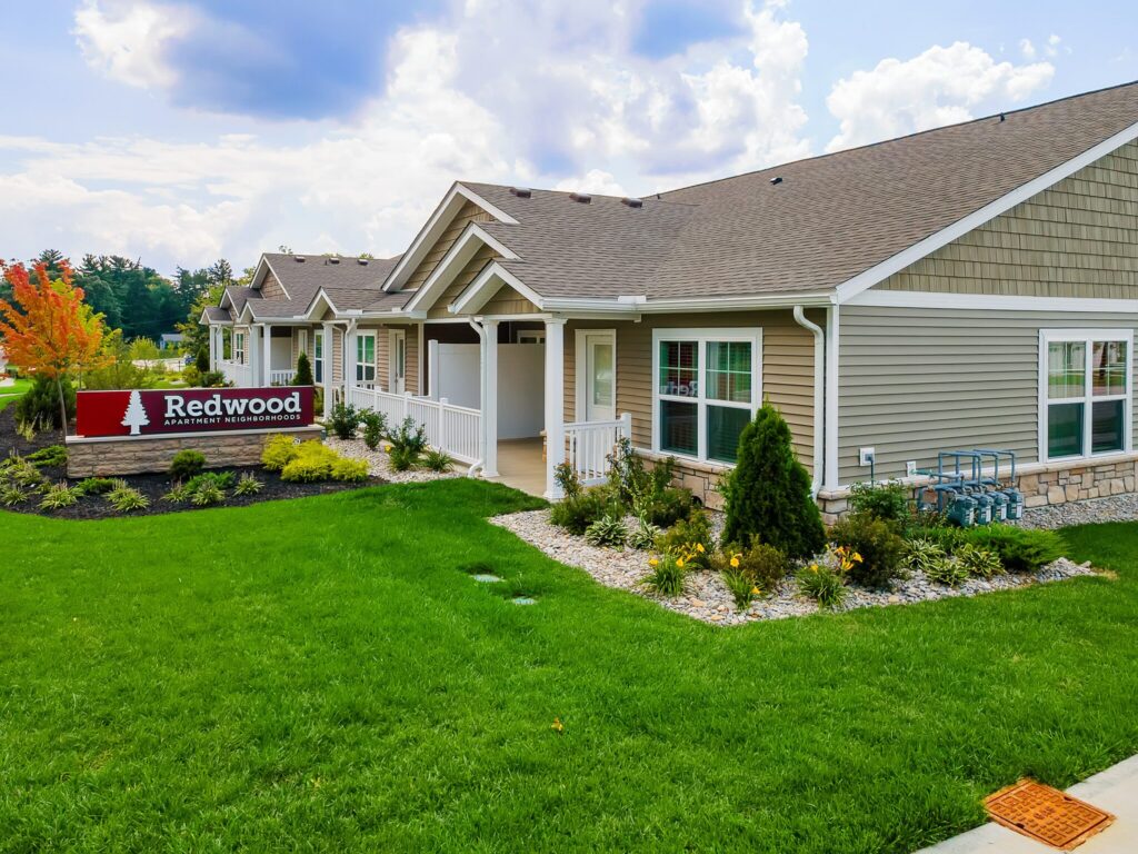 Single-story apartment units with beige siding and manicured landscaping. A red sign in front reads "Redwood Apartment Neighborhood." Cloudy sky in the background.