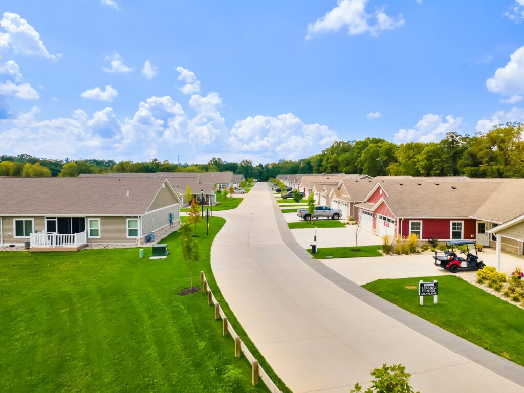 A suburban neighborhood street with single-story houses, green lawns, driveways, and a partly cloudy sky.