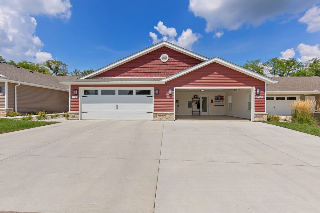 Single-story house with a red exterior, white trim, and a double garage; one garage door is open, revealing the interior. Large concrete driveway in front.