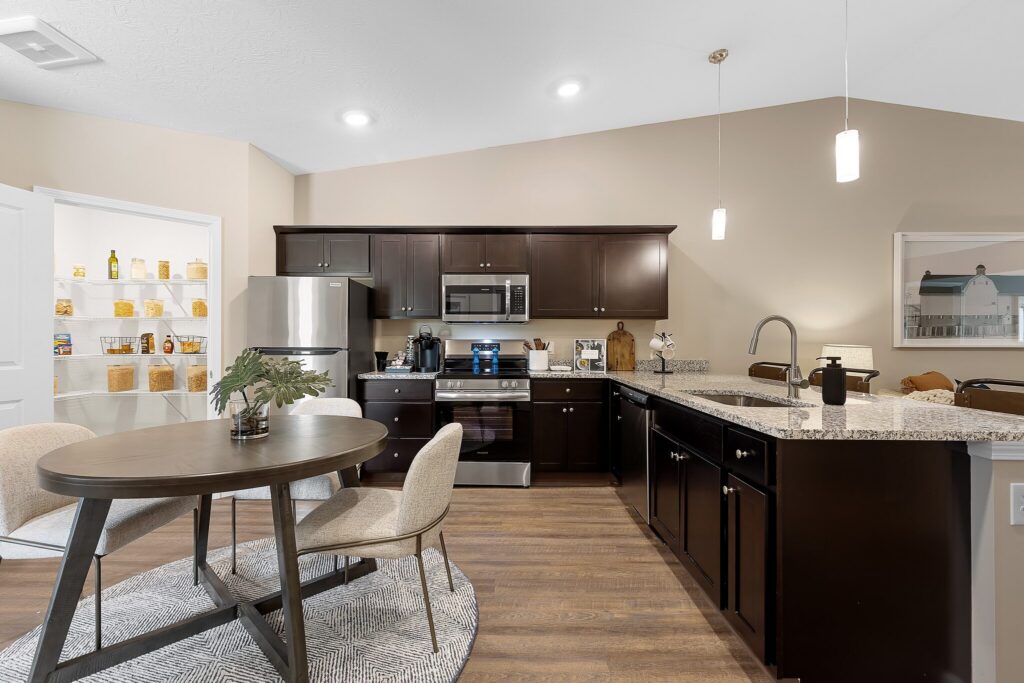 Modern kitchen with dark cabinets, stainless steel appliances, granite countertops, and a round dining table with two chairs; pantry with shelves visible in the background.