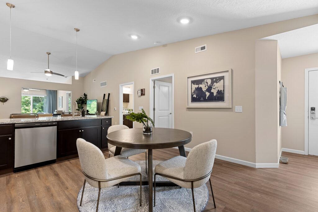Modern dining area with round table and four chairs on a rug, adjacent to a kitchen with dark cabinets and stainless steel appliances.