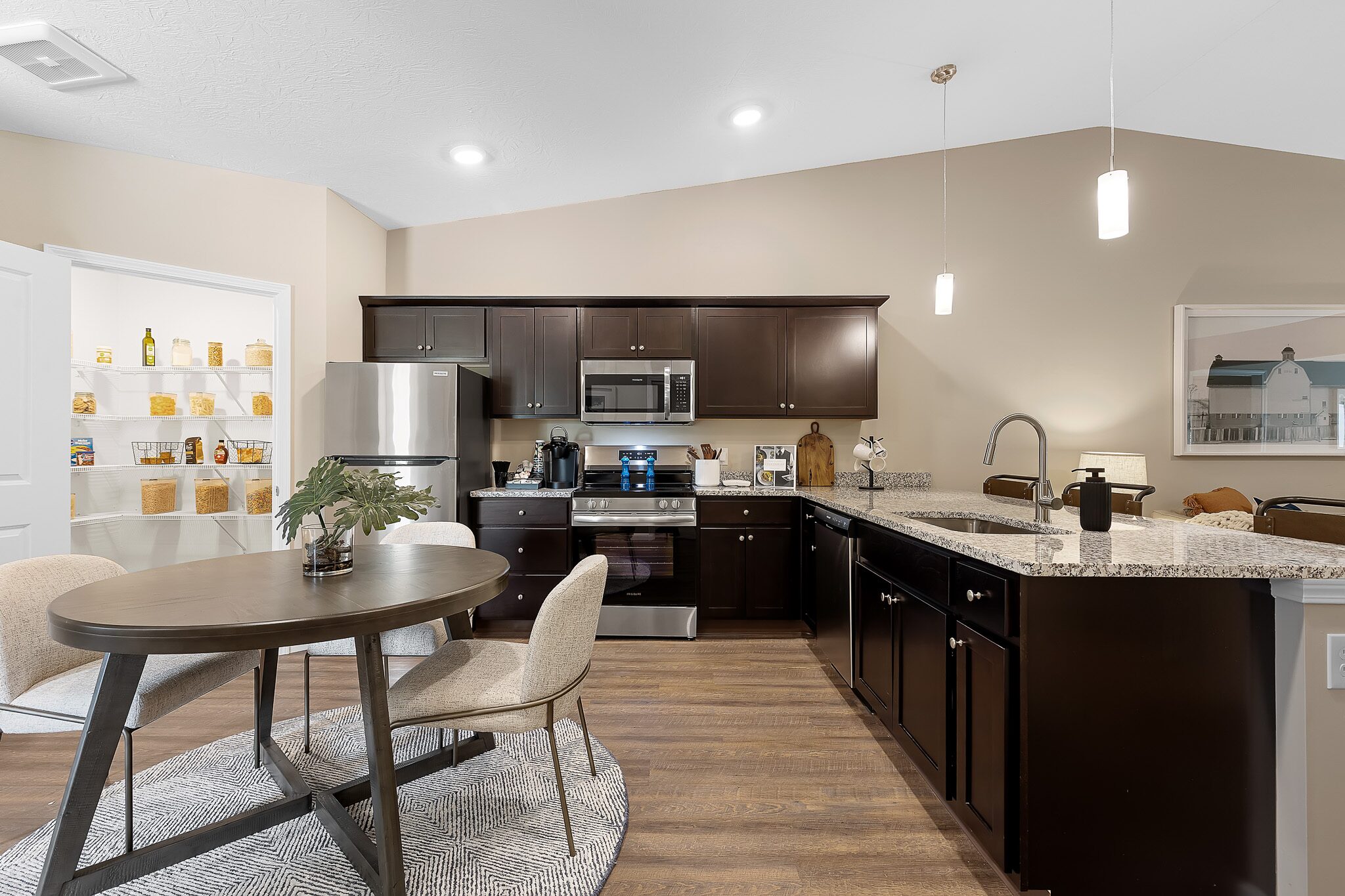 Modern kitchen with dark cabinets, stainless steel appliances, granite countertops, and a round dining table with two chairs; pantry with shelves visible in the background.