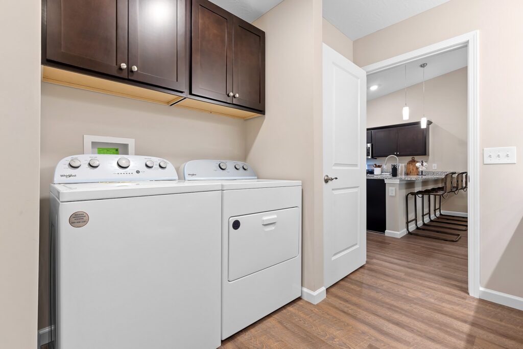 A laundry room with a white washer and dryer, dark wood cabinets above, beige walls, and an open door leading to a kitchen with bar stools and pendant lighting.