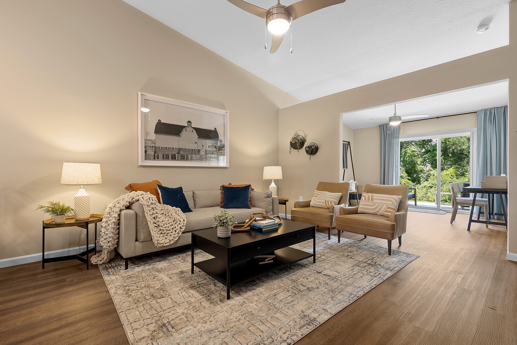 A modern living room with a sofa, two armchairs, a coffee table, and lamps, featuring wood flooring and large windows opening to a dining area with outdoor views.