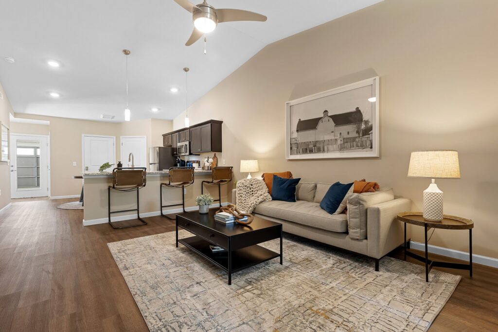 Modern open-concept living room and kitchen with neutral walls, wood floors, a beige sofa, bar stools at the counter, and a large barn artwork above the couch.