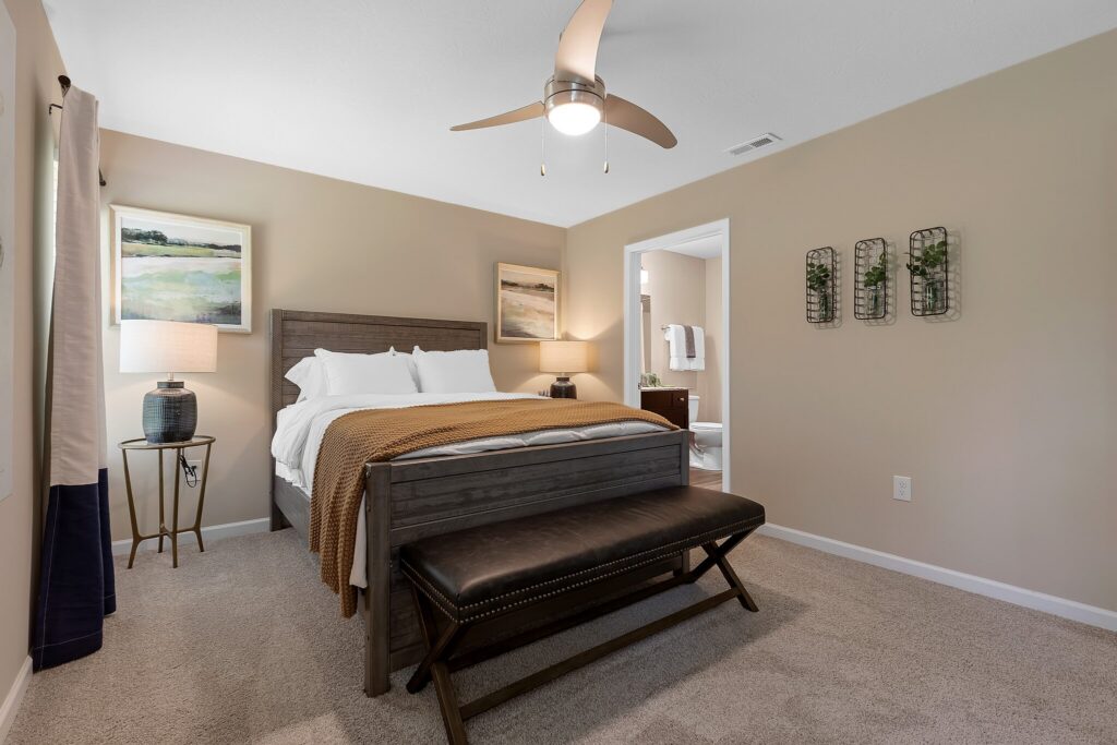 A neatly made bed with white and brown bedding is centered in a beige bedroom, featuring a ceiling fan, side table with lamp, bench, and decor on the walls.