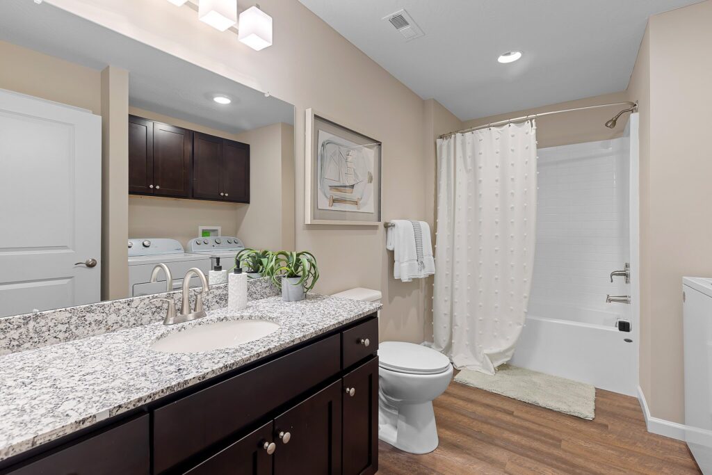 Modern bathroom with granite countertop, dark wood cabinets, toilet, bathtub with white shower curtain, wall art, towel rack, plants, and view of laundry machines in adjacent room.