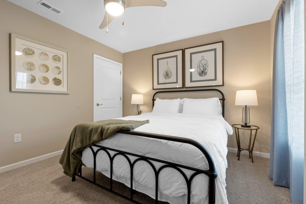 A neatly made bed with white bedding, two bedside lamps, framed artwork above the headboard, and a green blanket in a beige-walled bedroom.