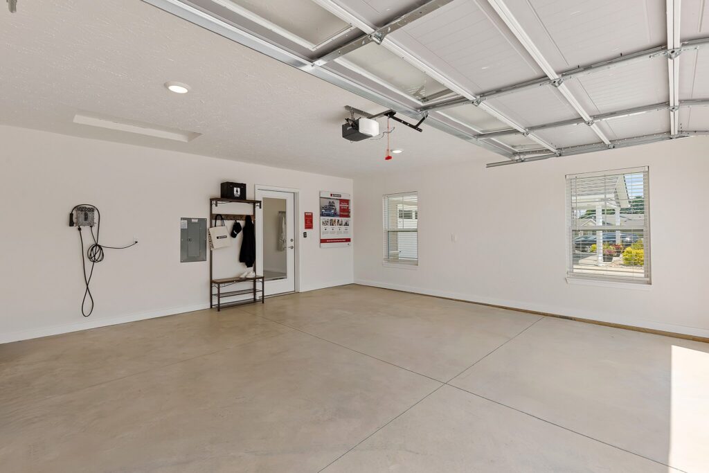 Spacious, clean garage with light-colored walls, concrete floor, ceiling-mounted garage door opener, windows, wall utilities, and a small shelving unit.