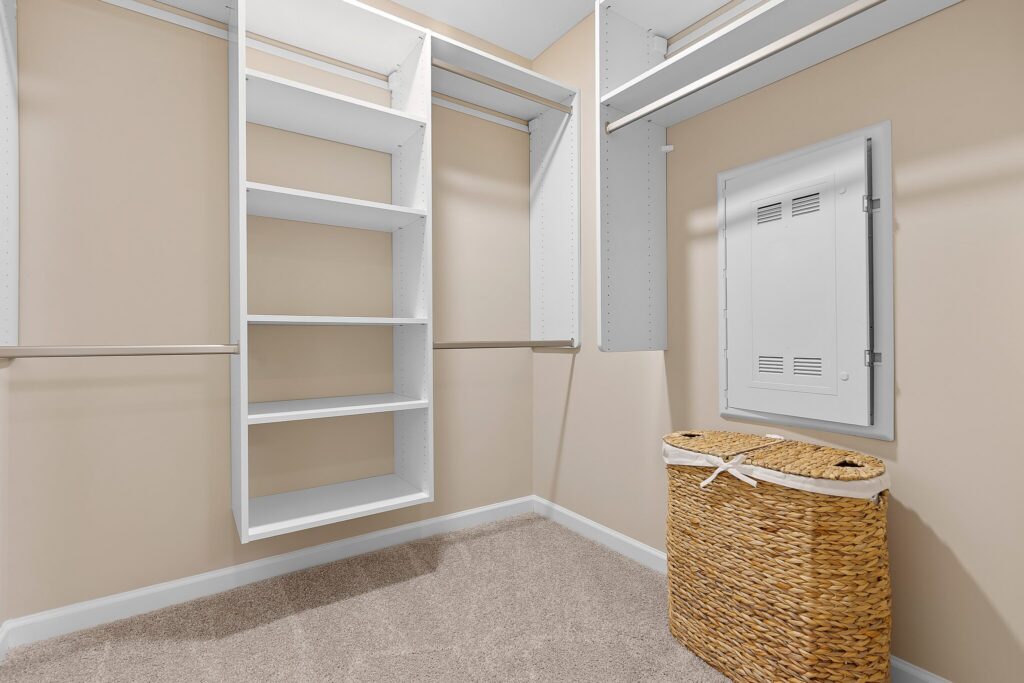 Empty walk-in closet with white shelving units, beige walls, carpeted floor, electrical panel on the wall, and a woven laundry basket in the corner.