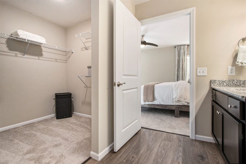 Open door reveals a walk-in closet with shelves and a laundry basket, adjacent to a bathroom vanity and a carpeted bedroom with a bed and ceiling fan.