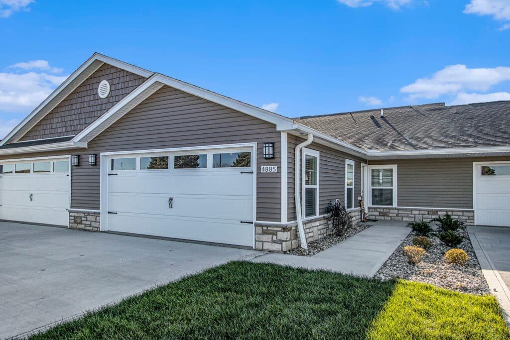 Single-story townhouse with brown siding, white trim, and a double garage. Concrete driveway and walkway, with minimal landscaping and green grass in front.