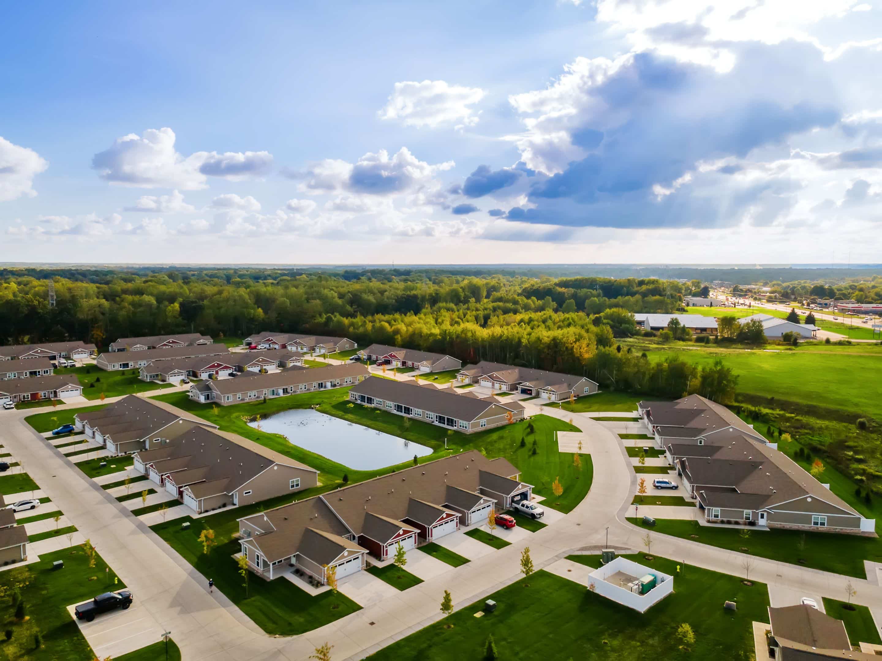 Aerial view of a suburban neighborhood with single-story homes, curved streets, green lawns, and a small pond, surrounded by trees under a partly cloudy sky.