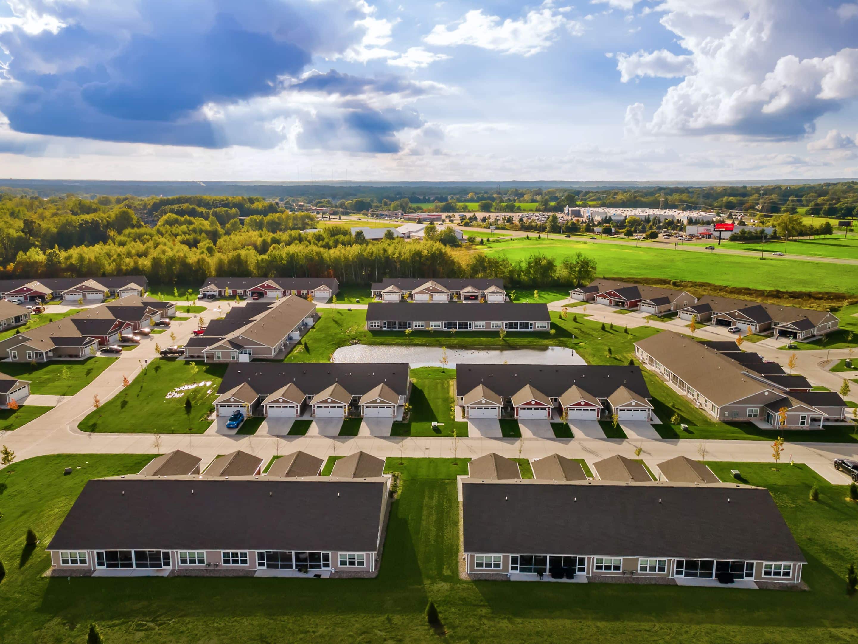 Aerial view of a suburban neighborhood with rows of single-story houses, green lawns, trees, and a partly cloudy sky.