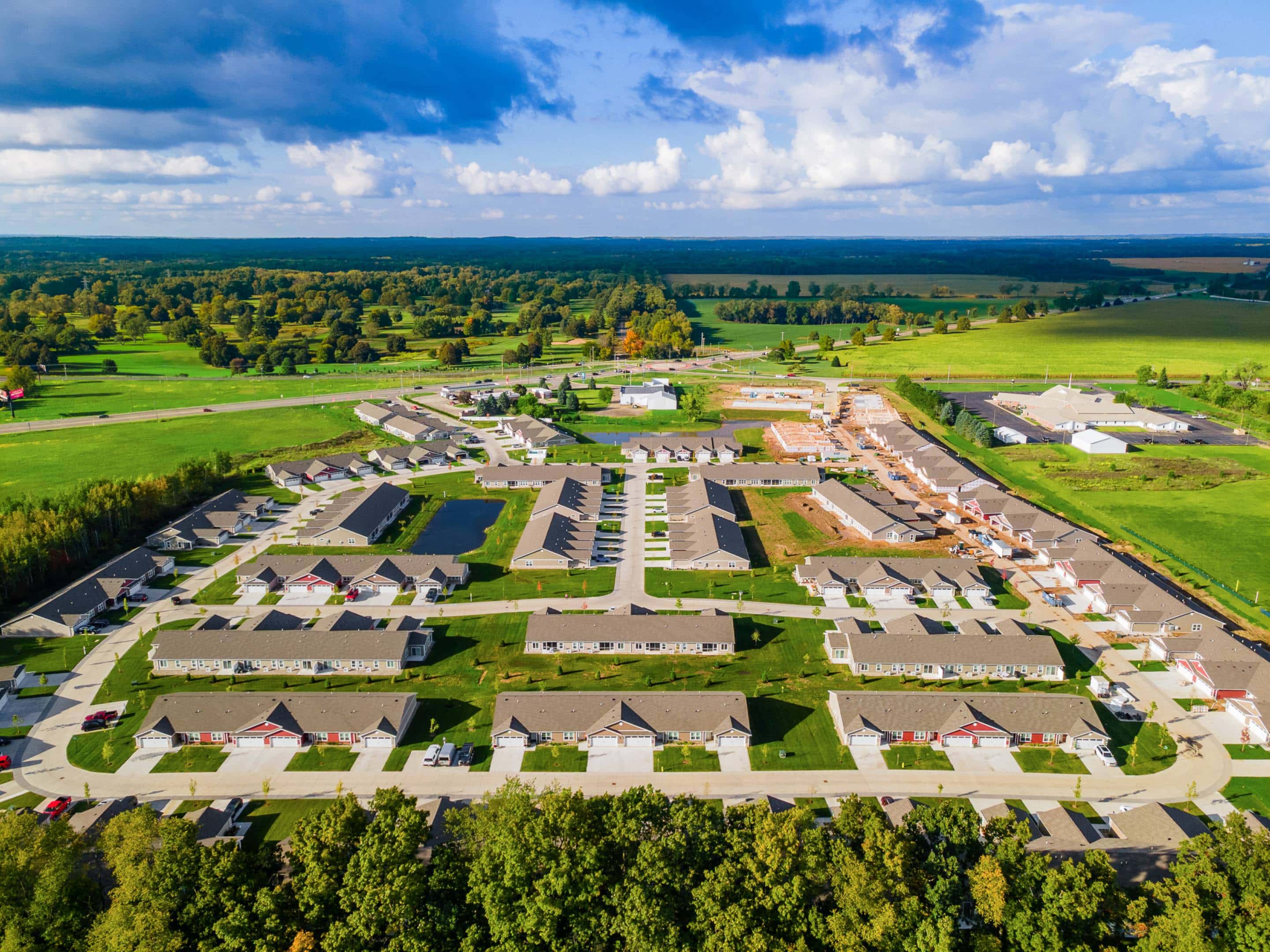 Aerial view of a suburban residential neighborhood with rows of houses, roads, green lawns, and surrounding open fields under a partly cloudy sky.