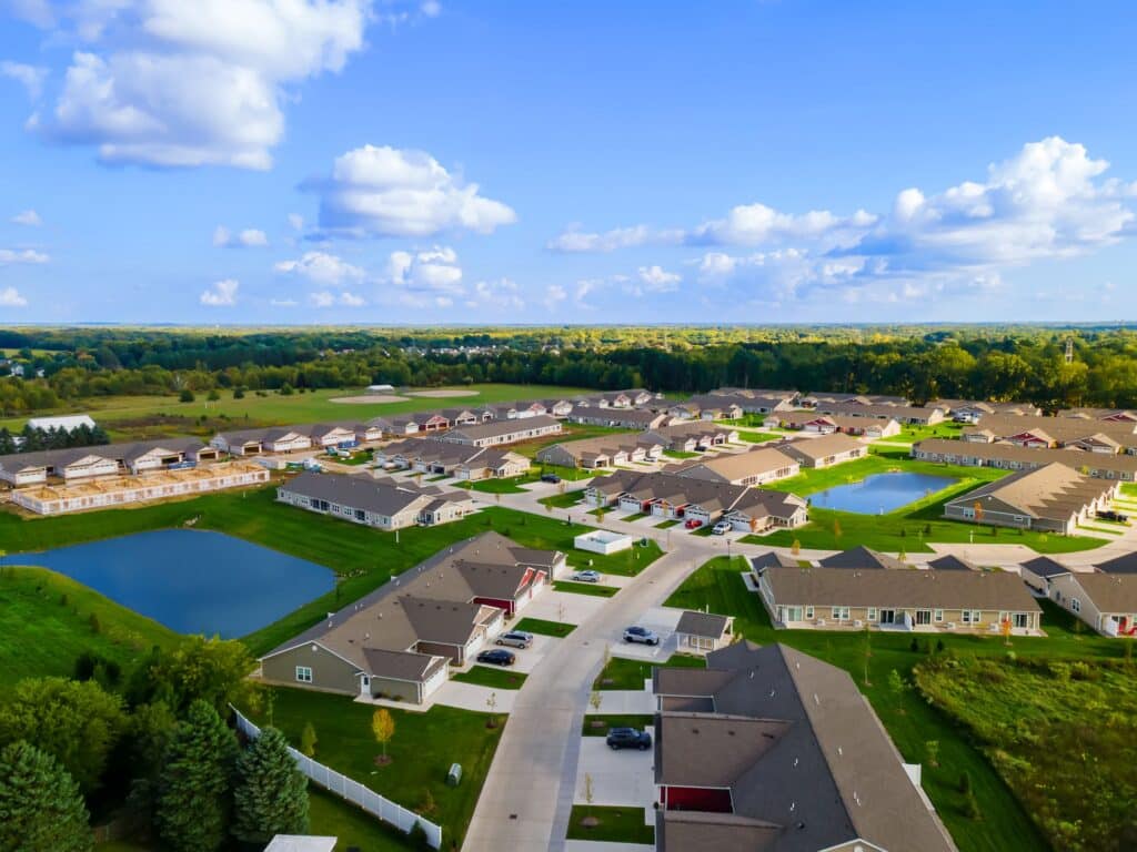 Aerial view of a suburban neighborhood with rows of houses, streets, green lawns, small ponds, and surrounding trees under a blue sky with clouds.