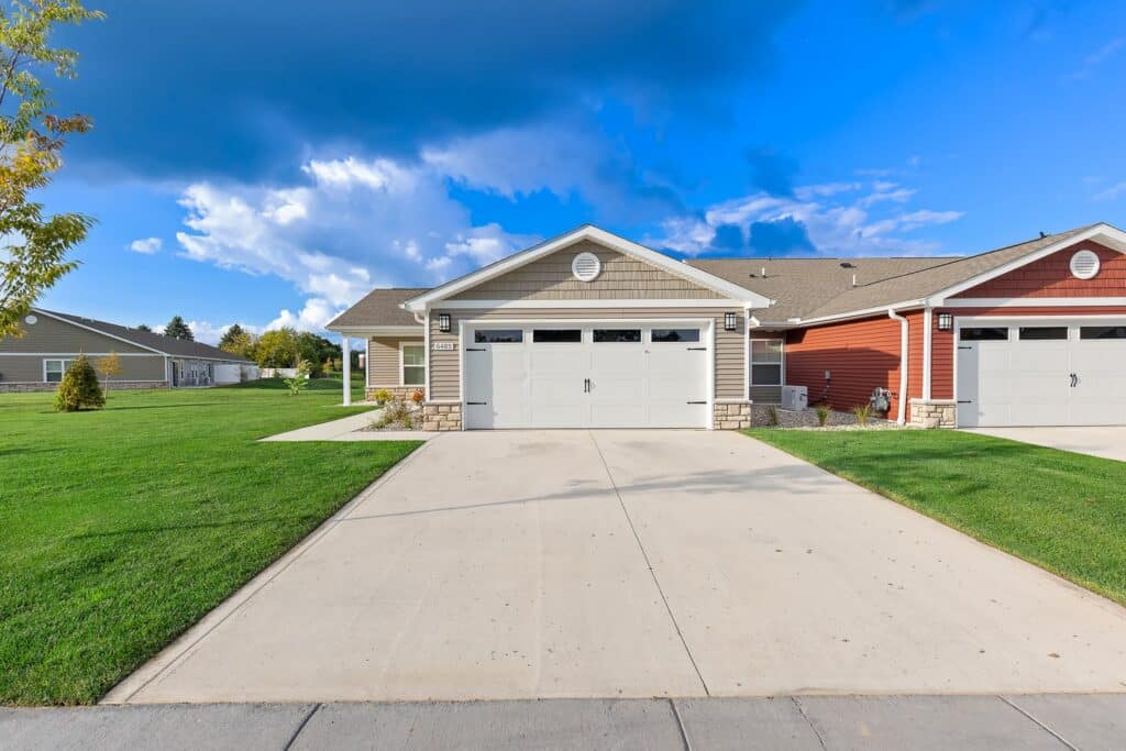 Single-story house with beige siding, white garage door, and concrete driveway, adjacent to a similar red house, on a well-manicured lawn under a partly cloudy sky.