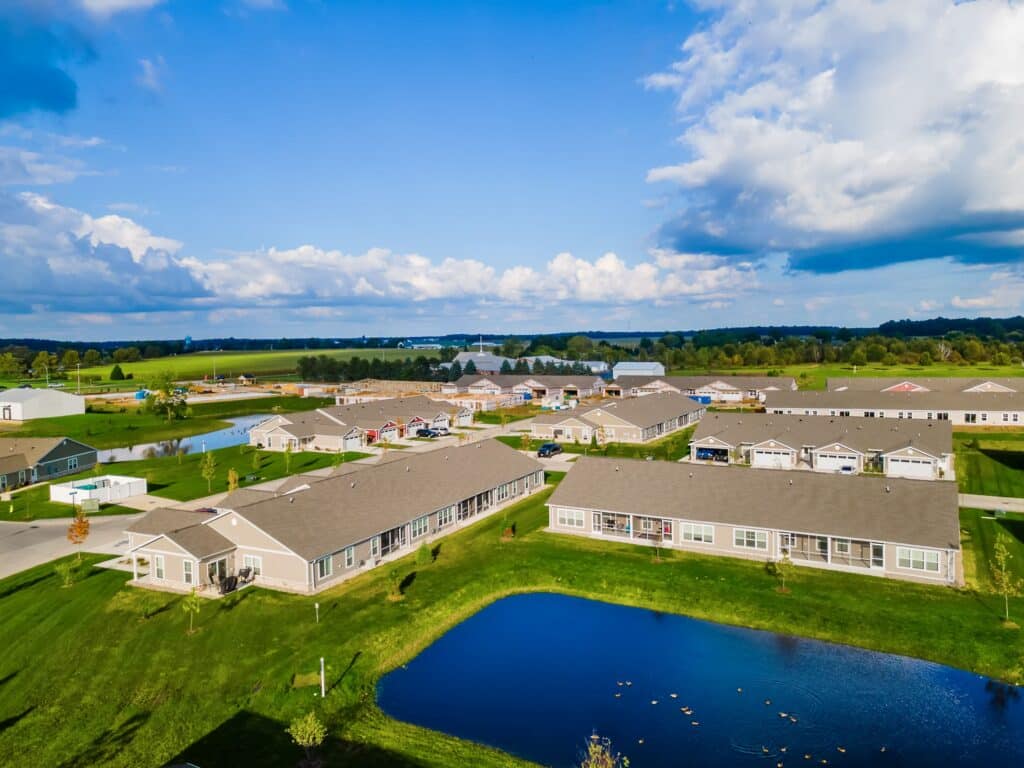 Aerial view of a residential neighborhood with single-story houses, green lawns, a small pond, and open fields in the background under a partly cloudy sky.