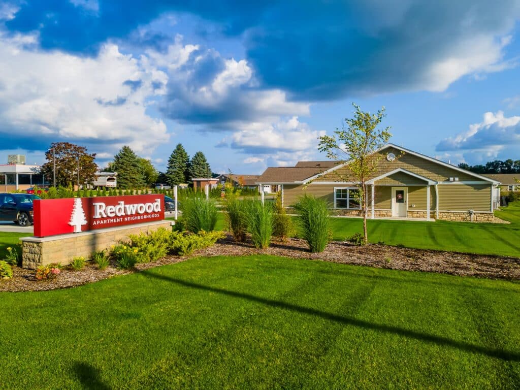 A single-story apartment building with a well-kept lawn and a red sign reading "Redwood Apartment Neighborhoods" in front.