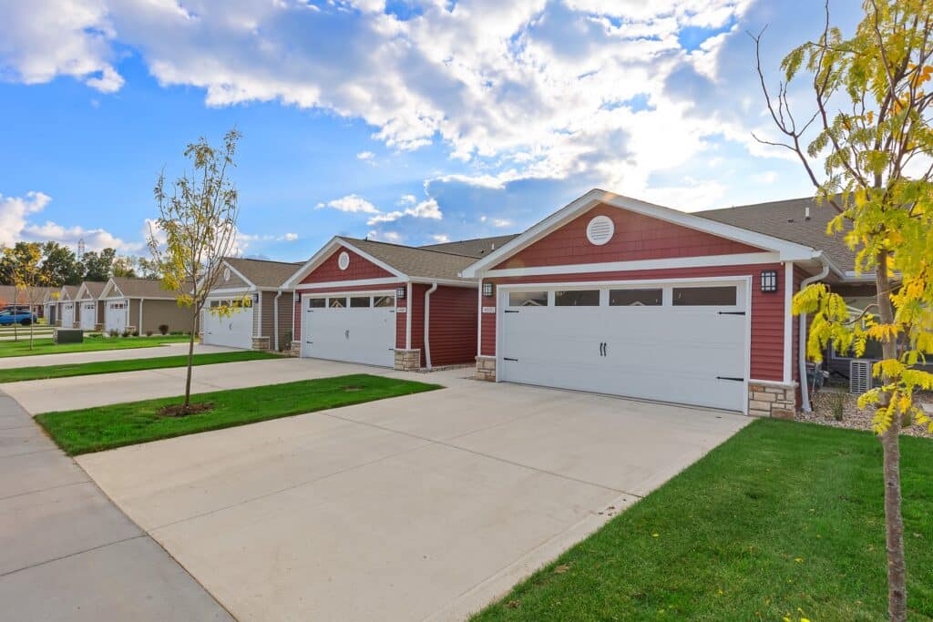 Row of attached single-story houses with red and beige siding, double garages, and neatly maintained lawns on a sunny day.