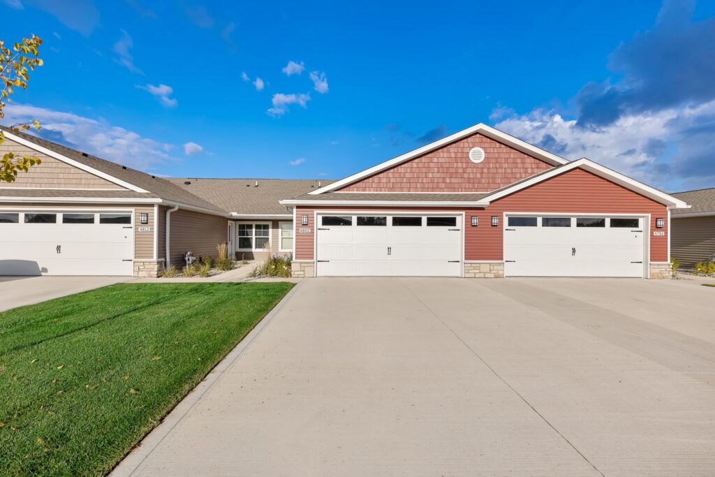 Duplex building with two connected units, each with a double garage, concrete driveway, red and beige siding, and manicured lawn under a blue sky.
