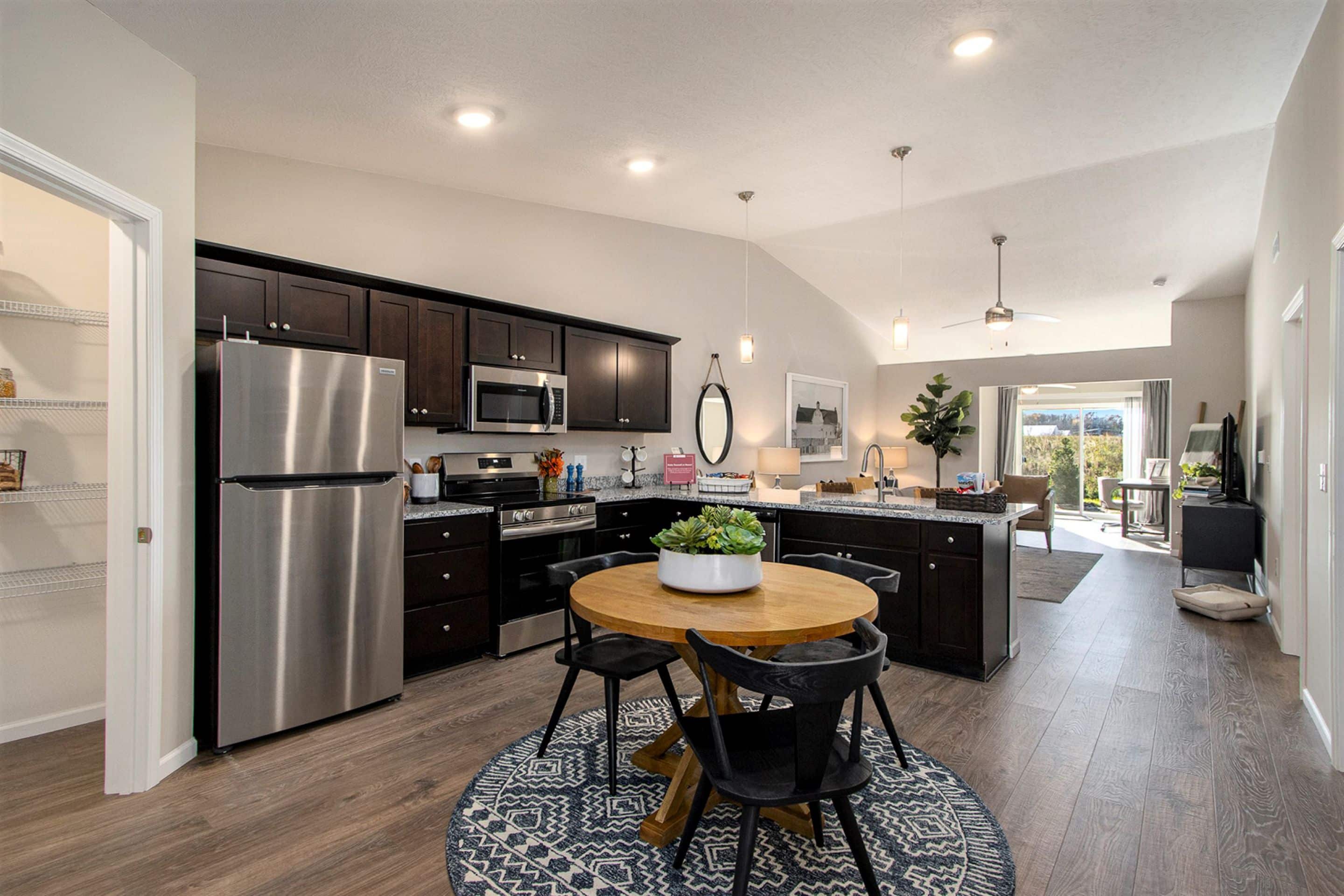 Modern open-concept kitchen and dining area with dark cabinets, stainless steel appliances, round table, and living room in the background.