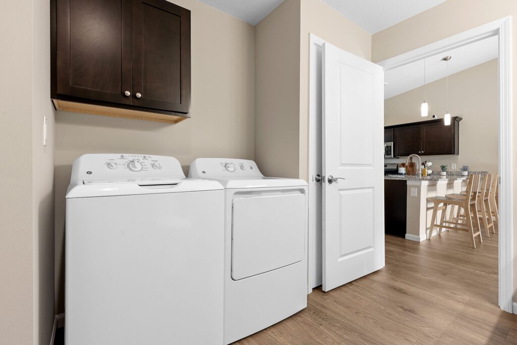 A laundry room with a white washing machine and dryer side by side, a dark wood wall cabinet above, and an open door leading to a kitchen and dining area.