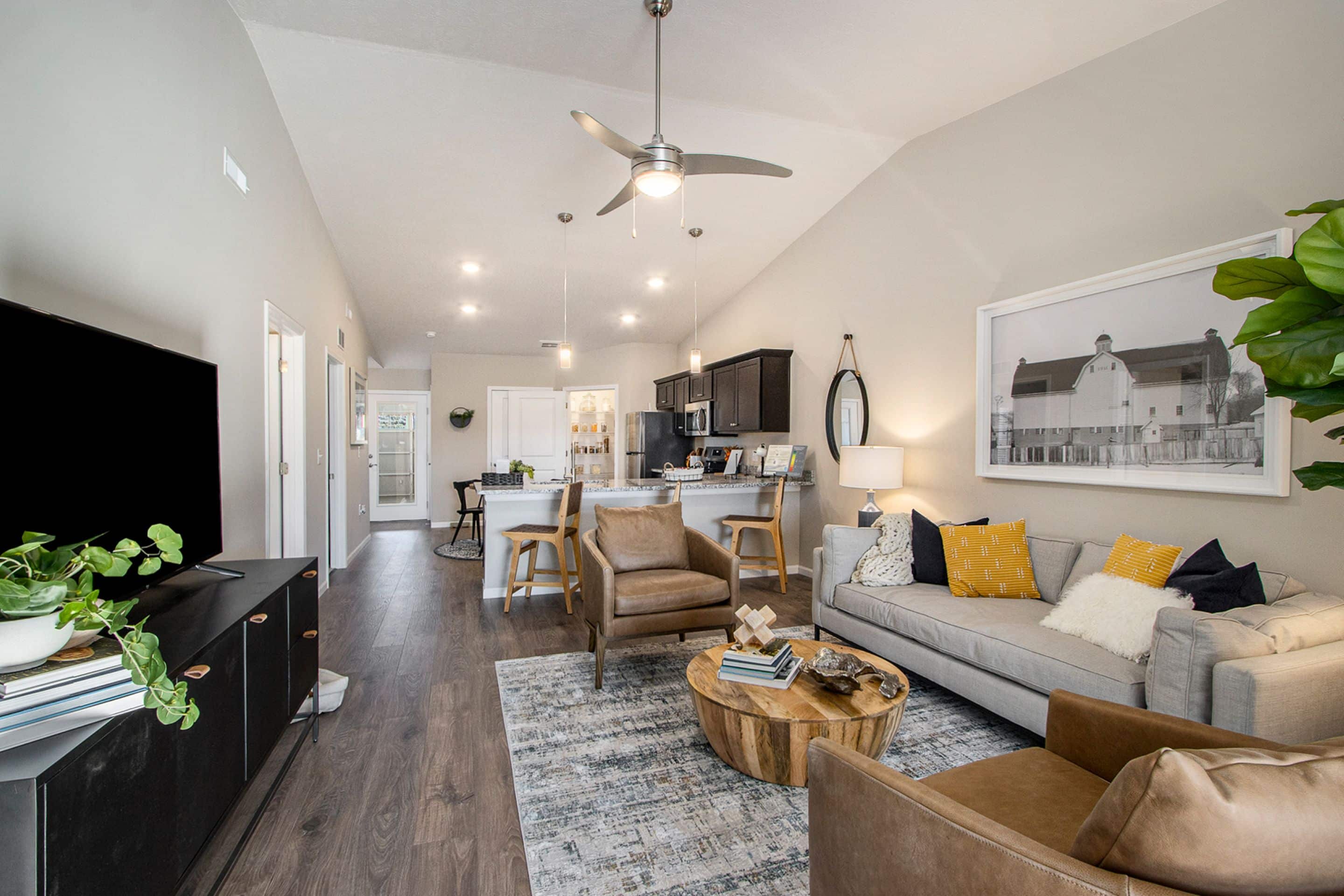 Modern living room with neutral tones, a gray sofa, two brown leather chairs, a round wooden coffee table, large wall art, and an open kitchen in the background.