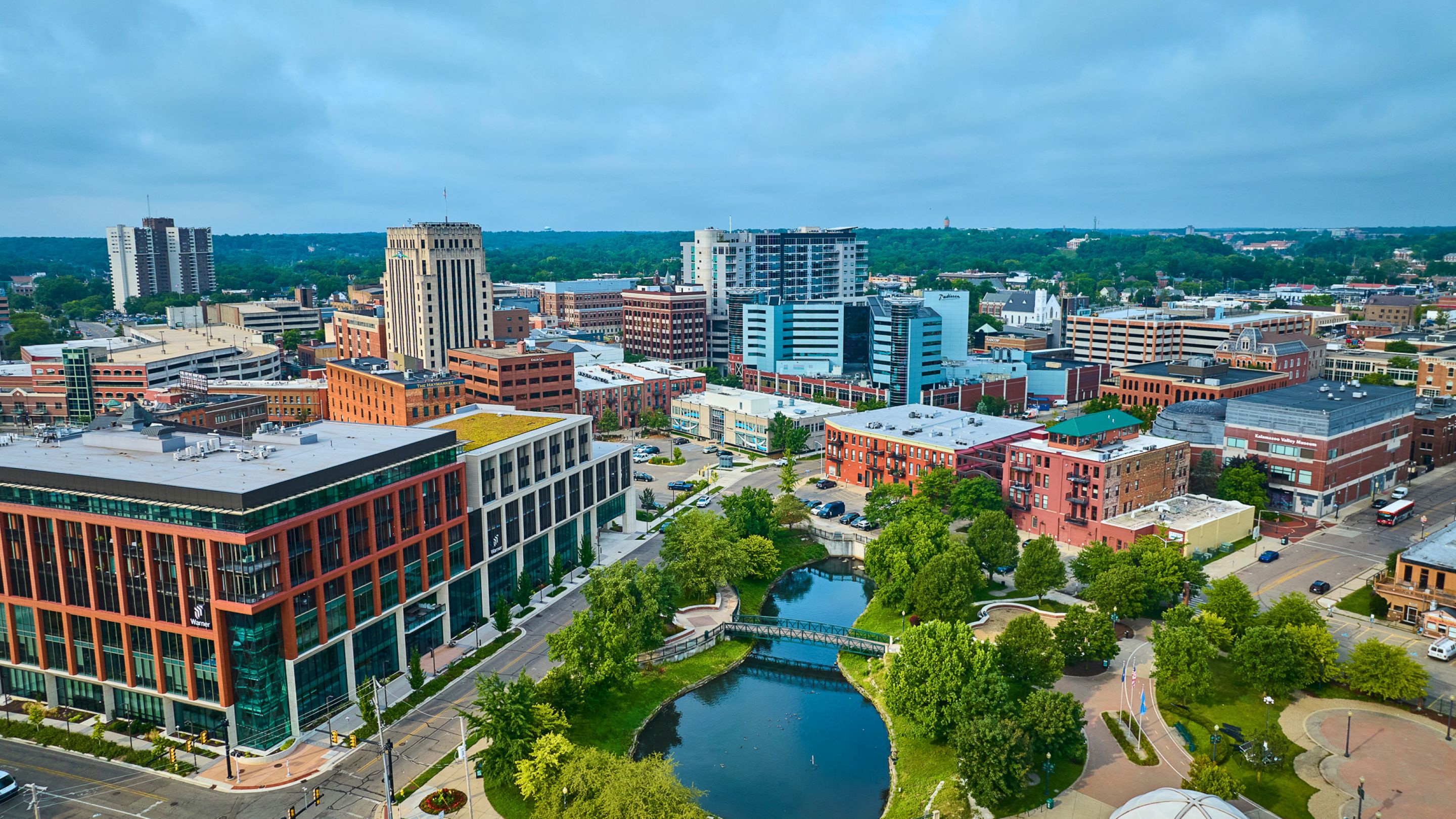 Aerial view of a cityscape with modern and historic buildings, a small park, pond, and trees in the foreground under a cloudy sky.